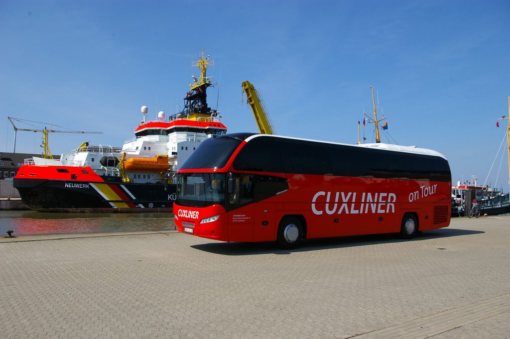 Ein roter CUXLINER Reisebus steht am Hafen mit dem Seenotrettungskreuzer NEUWERK im Hintergrund. CUXLINER "on Tour" bietet Busreisen und Schiffsreisen ab Cuxhaven an.