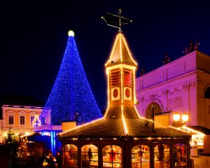 Eine bezaubernde Weihnachtsmarktszene bei Nacht, erleuchtet von warmen goldenen Lichtern. Ein großer, blau beleuchteter Weihnachtsbaum steht hinter einem malerischen Holzpavillon, geschmückt mit funkelnden Lichtern und festlichen Dekorationen. Die Szenerie ruft eine gemütliche und zauberhafte Weihnachtsstimmung hervor.