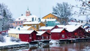Winterliche Ansicht von Porvoo, Finnland, mit farbenfrohen Holzhäusern am Flussufer unter einer Schneedecke. Der Turm der alten Stadtkirche ragt im Hintergrund hervor.