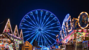 Das ist ein Foto vom Weihnachtsmarkt in Rostock mit einem blau leuchteten Riesenrad im Bildmittelpunkt. Es ist abends und dunkel. Links und rechts sind Dächer von weihnachtlich geschmückten Buden und ein Karussell zu sehen.
