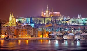Nächtliches Stadtbild von Prag im Winter mit schneebedeckten Dächern und der beleuchteten Karlsbrücke. Die Prager Burg und der Veitsdom erheben sich majestätisch im Hintergrund und spiegeln sich in der Moldau.