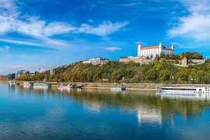 Panaramaansicht von Bratislava mit der Burg als zentrales Motiv. Am Ufer der Donau liegen mehrere Flusskreuzfahrtschiffe.