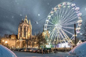 Winterliche Erlebnisreisen in Erfurth: Ein beleuchtetes Riesenrad vor einer gotischen Kirche unter Schneefall. Die Lichter des Riesenrads spiegeln sich im Schnee, eine zauberhafte Szene.
