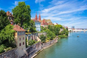 Eine hohe Mauer verläuft direkt Am Rhein entlang. Ein Turm ist in die Mauer eingelassen und überblickt das Wasser. Hinter der begrünten Anlage sind zahlreiche historische Gebäude zu sehen, zwei Türme überragen diese.