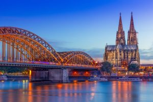 Eine am Abend warm beleuchtete Hohenzollernbrücke überspannt den Rhein. Auf der anderen Uferseite wird der Kölner Dom angestrahlt. Mehrere Fahrgastschiffe liegen am Ufer.