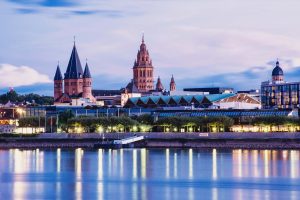 Abendlicher Blick auf Mainz am Rhein. Der Mainzer Dom prägt die beleuchtete Skyline. In den Rhein ragt ein Schiffsanleger.