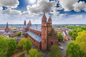 Erhöhte Ansicht des Wormser Doms. Markante Türme prägen das Bauwerk. Umgeben ist der Dom von vielen grünen Bäumen und städtischen Gebäuden.