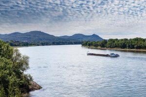 Ein Frachtschiff fährt den Rhein bei Bonn entlang. Die Ufer sind von Bäumen gesäumt, im Hintergrund sind einige Berge sichtbar.