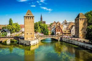 An einem Fluss stehen zwei Türme, einer am rechten Ufer, der andere auf einer Flussinsel in der Mitte. Zwei kleine Bogenbrücken verbinden die Ufer mit der Flussinsel. Im Hintergrund sind historische Gebäude zu sehen, einige mit Fachwerk.