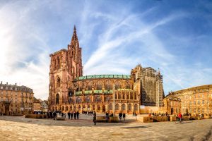 Gesamtansicht des Straßburger Münsters. Im Vordergrund ein großer Platz, auf dem zahlreiche Menschen verweilen. Historische Gebäude umgeben das Münster.