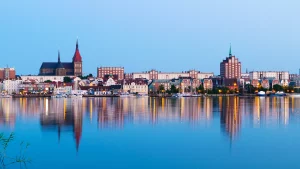 Panorama von der Stadt Rostock. Im Vordergrund ist der Fluss Warnow, in dem sich die Gebäude der Stadt entlang des Ufers spiegeln.