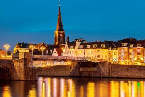 Nächtliche Ansicht von Maastricht, Niederlande, mit beleuchteten Häusern, Brücke und Kirchturm, die sich im Wasser spiegeln. Warme, goldene Lichter erhellen die Szene unter einem tiefblauen Himmel.