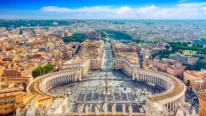 Panorama des Petersplatzes in Vatikanstadt, Rom, mit Blick auf die Kolonnaden, den Obelisken und die weitläufige Stadtlandschaft unter blauem Himmel.