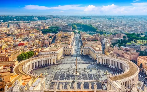 Panorama des Petersplatzes in Vatikanstadt, Rom, mit Blick auf die Kolonnaden, den Obelisken und die weitläufige Stadtlandschaft unter blauem Himmel.