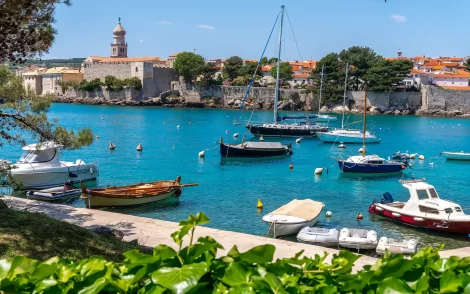 Kroatische Küstenstadt Krk mit türkisfarbenem Wasser und zahlreichen Booten vor der historischen Stadtmauer mit Kirchturm. Einladende Szene aus dem Reisekatalog 2026.