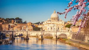 Rom-Panorama mit Blick auf den Petersdom über den Tiber und die Engelsbrücke im Sonnenlicht. Blühende Zweige im Vordergrund. Reisekatalog 2026.