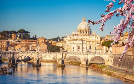 Rom-Panorama mit Blick auf den Petersdom über den Tiber und die Engelsbrücke im Sonnenlicht. Blühende Zweige im Vordergrund.