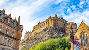 Edinburgh Castle thront auf einem Felsen über der Stadt, umgeben von historischen Gebäuden und üppigem Grün unter einem hellblauen Himmel.