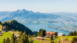 Panoramablick auf den Vierwaldstättersee und die Schweizer Alpen vom Berg Rigi. Eine rote Zahnradbahn schlängelt sich durch grüne Wiesen. Der neue Reisekatalog 2026 lädt zur Entdeckung ein!