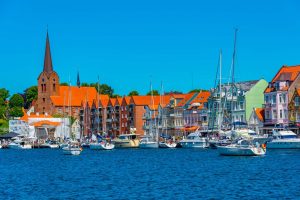 Hafenansicht von Schleswig, Deutschland, mit bunten Häusern, Segelbooten und der St. Petri Kirche unter blauem Himmel. Lebendige Szene an der Schlei.