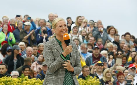 Blonde Moderatorin mit orangefarbenem Mikrofon spricht vor einer Menschenmenge im Freien. Sie trägt ein kariertes Jackett über einem grünen Kleid. Publikum im Hintergrund.