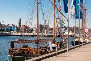 Historisches Segelschiff im Hafen von Kiel, Deutschland. Ein Mädchen fotografiert die Takelage. Im Hintergrund die Stadtkulisse mit Kirchturm und modernen Gebäuden unter blauem Himmel.