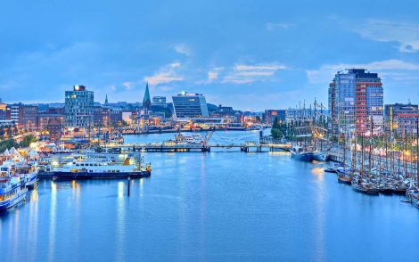 Hafenansicht von Kiel bei Dämmerung. Zahlreiche Segelschiffe und moderne Gebäude spiegeln sich im ruhigen Wasser. Die blaue Stunde taucht die Stadt in ein malerisches Licht.