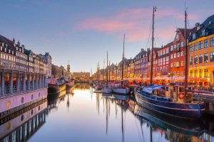 Farbenfrohe Häuserfassaden spiegeln sich im ruhigen Wasser des Nyhavn Kanals in Kopenhagen. Segelboote liegen vertäut, während das Abendlicht eine malerische Szene schafft.