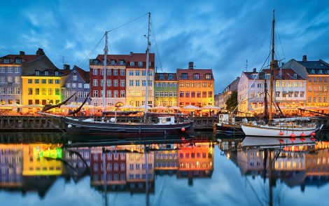 Farbenfrohe Häuserfassaden spiegeln sich im Wasser des Nyhavn in Kopenhagen. Segelboote liegen vor Anker, abends beleuchtet.