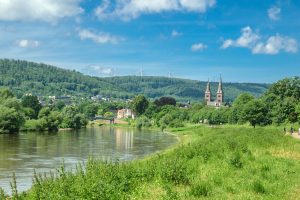 Weser-Flusslandschaft in Deutschland mit grünen Ufern, einer Stadt im Hintergrund und Windrädern auf einem bewaldeten Hügel. Zwei Kirchtürme ragen über die Stadt hinaus.