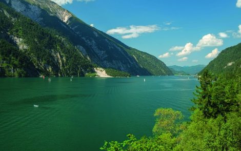 Türkisblauer Achensee, eingebettet zwischen bewaldeten Bergen unter blauem Himmel mit Wolken. Segelboote und ein kleines Boot auf dem See.