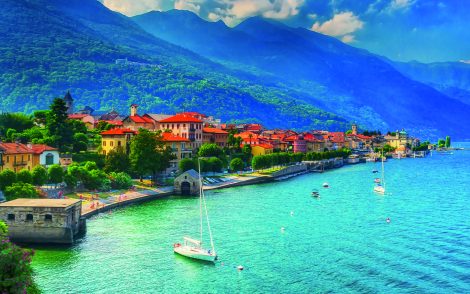 Malerische Küstenstadt in Italien mitAdobeStock_218548211 bunten Häusern am Comer See, umgeben von grünen Bergen unter blauem Himmel. Segelboote ankern im türkisfarbenen Wasser.