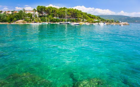 Türkisblaues Meerwasser vor einer grünen, bewaldeten Küste mit weißen Häusern und Segelbooten. Sonniger Tag an der Adria, Kroatien.