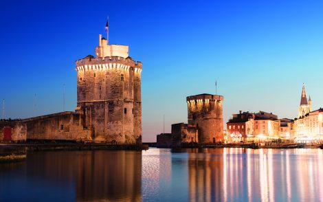 Zwei beleuchtete mittelalterliche Türme in La Rochelle, Frankreich, spiegeln sich im ruhigen Wasser des Hafens unter einem tiefblauen Abendhimmel. Die Stadtkulisse mit Kirchturm ist im Hintergrund zu sehen.