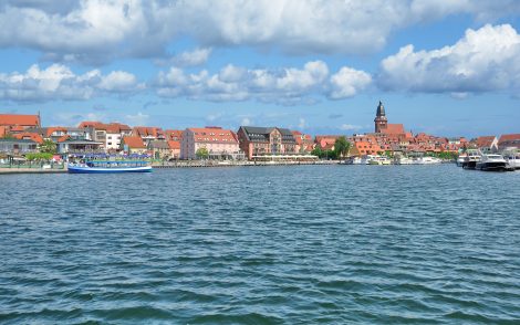 Blick über das Wasser auf Waren (Müritz) mit bunten Häusern, rotem Kirchturm und blauen Himmel. Boote liegen im Hafen.