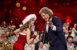 André Rieu spielt Geige bei einem Adventskonzert in Maastricht. Eine junge Frau im roten Weihnachtskleid und Nikolausmütze tanzt begeistert neben ihm, vor einem festlichen roten Hintergrund.