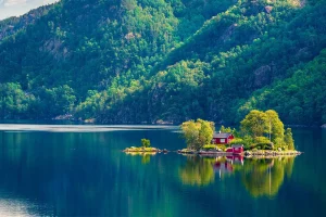 Idyllische norwegische Landschaft: Rotes Haus auf einer kleinen Insel in einem ruhigen Fjord, umgeben von grünen Bäumen und Bergen. Spiegelung im klaren, blauen Wasser.