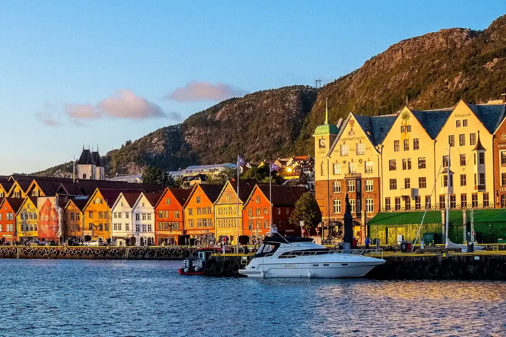 Farbenfrohe Häuserfassaden in Bryggen, Bergen, Norwegen, spiegeln sich im Hafenwasser. Ein Boot liegt vor der historischen UNESCO-Weltkulturerbestätte, mit Bergen im Hintergrund.