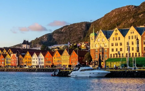 Farbenfrohe Häuserfassaden in Bryggen, Bergen, Norwegen, spiegeln sich im Hafenwasser. Ein Boot liegt vor der historischen UNESCO-Weltkulturerbestätte, mit Bergen im Hintergrund.