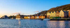 Farbenfrohe Häuserfassaden am Hafen von Bergen, Norwegen, im goldenen Abendlicht. Ein Segelschiff und moderne Boote liegen im ruhigen Wasser. Im Hintergrund erhebt sich ein bewaldeter Hügel.