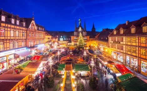 Weihnachtsmarkt in Werningerode bei Nacht: Blick auf den festlich beleuchteten Marktplatz mit Buden, Weihnachtsbaum und Fachwerkhäusern. Menschen bummeln durch den Markt, genießen die Atmosphäre.