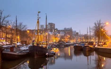 Winterliche Grachten in den Niederlanden bei Dämmerung. Festlich geschmückte Boote liegen im Wasser, die Lichter spiegeln sich im Kanal. Verschneite Dächer und Bäume im Hintergrund.