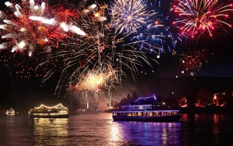 Feuerwerksspektakel über dem Wasser mit beleuchteten Ausflugsschiffen. Farbenprächtige Explosionen in Rot, Gold und Blau erhellen den Nachthimmel.