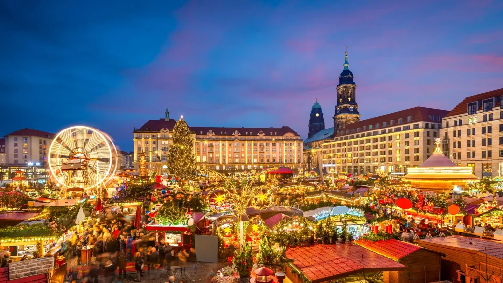 Dresdner Weihnachtsmarkt bei Nacht: Lebhaftes Treiben beim Striezelmarkt mit Lichterglanz, Riesenrad und festlich geschmücktem Weihnachtsbaum vor historischer Kulisse. Alles ist in warmes goldenes Licht gehüllt.