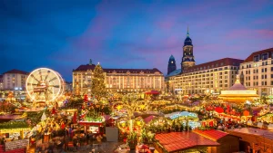 Dresdner Weihnachtsmarkt bei Nacht: Lebhaftes Treiben beim Striezelmarkt mit Lichterglanz, Riesenrad und festlich geschmücktem Weihnachtsbaum vor historischer Kulisse. Alles ist in warmes goldenes Licht gehüllt.