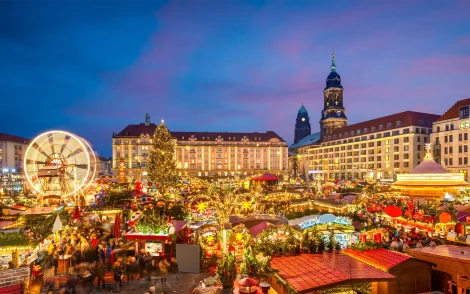 Dresdner Weihnachtsmarkt bei Nacht: Lebhaftes Treiben beim Striezelmarkt mit Lichterglanz, Riesenrad und festlich geschmücktem Weihnachtsbaum vor historischer Kulisse. Alles ist in warmes goldenes Licht gehüllt.
