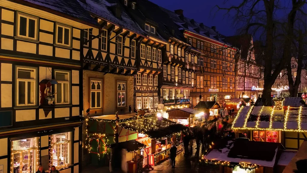 Marktplatz in Goslar bei Nacht, geschmückt mit Weihnachtsmarktständen und Lichterketten vor Fachwerkhäusern. Festliche Stimmung mit beleuchteten Buden und Menschen.