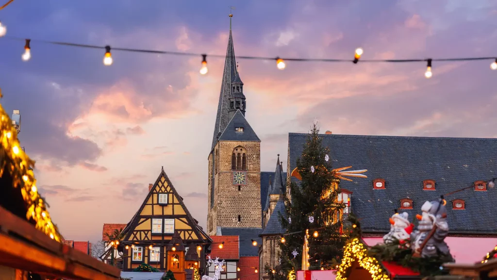 Abenddämmerung auf dem Weihnachtsmarkt in Quedlinburg. Im Hintergrund ragt der hohe Turm der historischen Kirche empor. Es sind geschmückte Buden und funkelnde Lichterketten angedeutet.