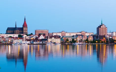 Das ist ein Foto von der Stadt Rostock. Im Vordergrund ist der Fluss Warnow, in dem sich die Gebäude der Stadt entlang des Ufers spiegeln.