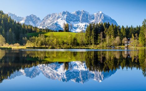 Spiegelglatter Kitzbuehel See reflektiert majestätische, schneebedeckte Berge und grüne Wälder. Idyllische Landschaft mit einem kleinen Haus auf einer Wiese. Perfekte Alpenkulisse.